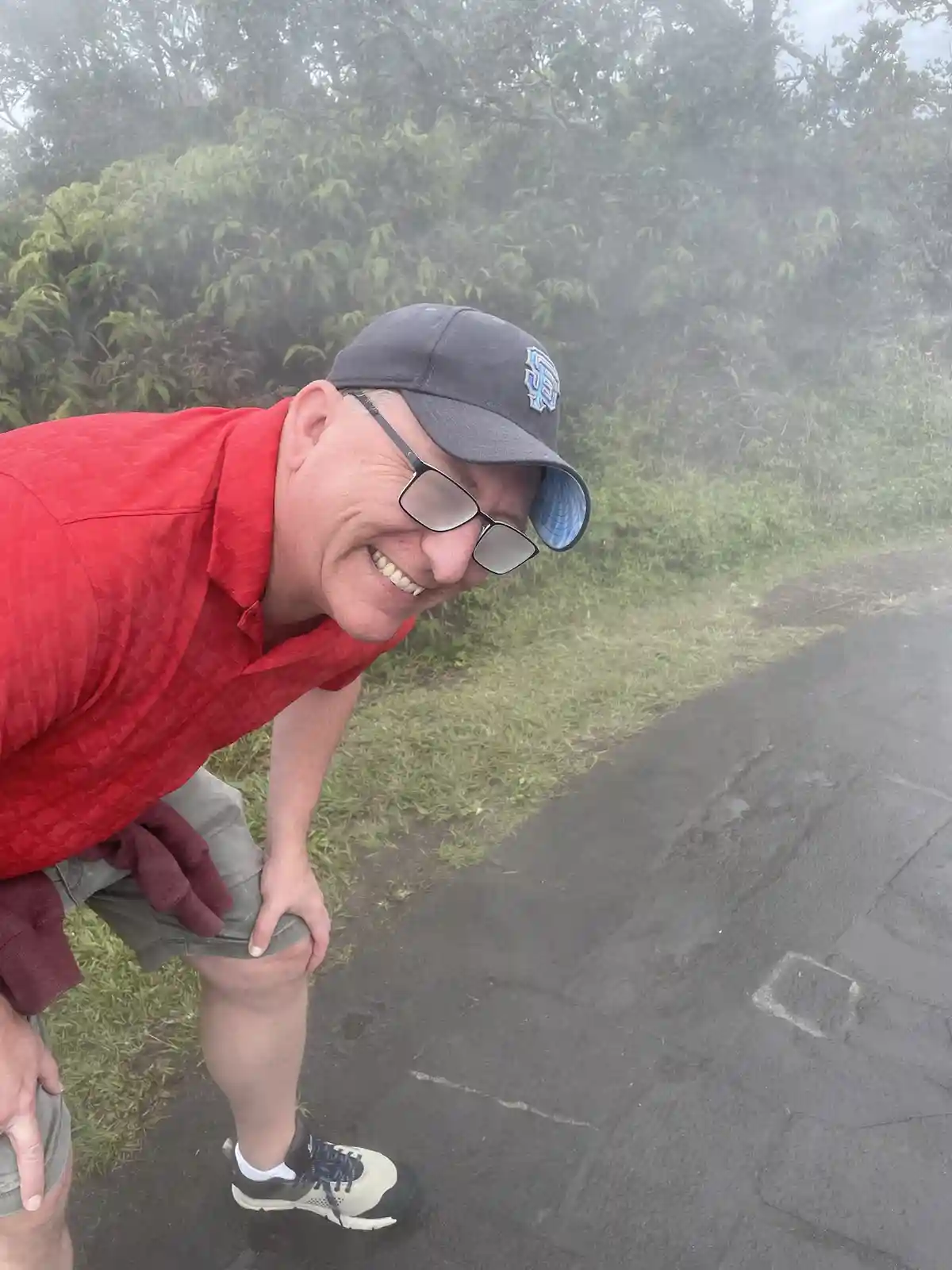 a man in a red shirt and grey cap is bent over on a misty path, smiling broadly while resting his hands on his knees. The…