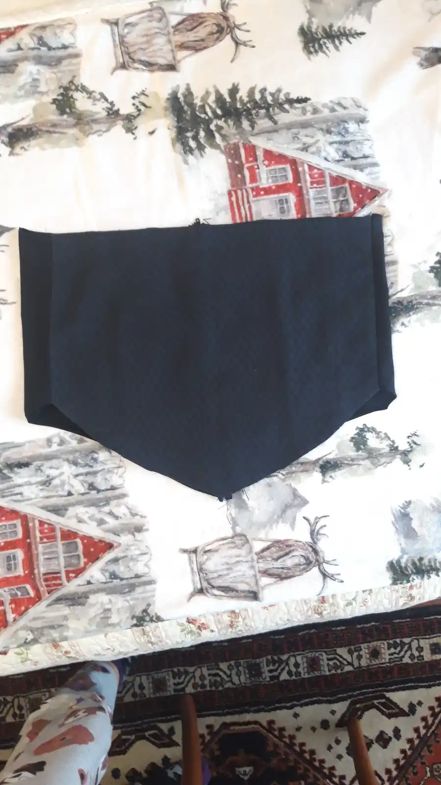 A dark blue, triangular-shaped skirt made from fabric lies flat on a quilt with a winter scene of red cabins and deer. The…