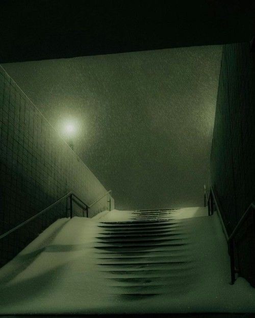 A dimly lit, snow-covered staircase at night, viewed from below, with a single overhead light casting a glow on the falling…