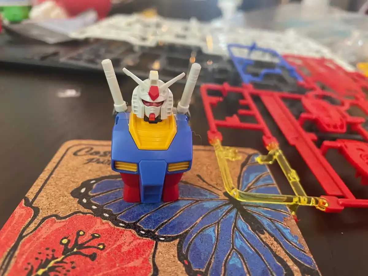 A small, assembled blue, yellow, and red plastic robot model, resembling a Gundam, stands upright on a cork coaster with a…