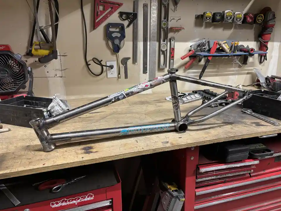 A custom-built, silver BMX bike frame with 'GRIZZLY' branding lies on a wooden workbench in a cluttered workshop. The…