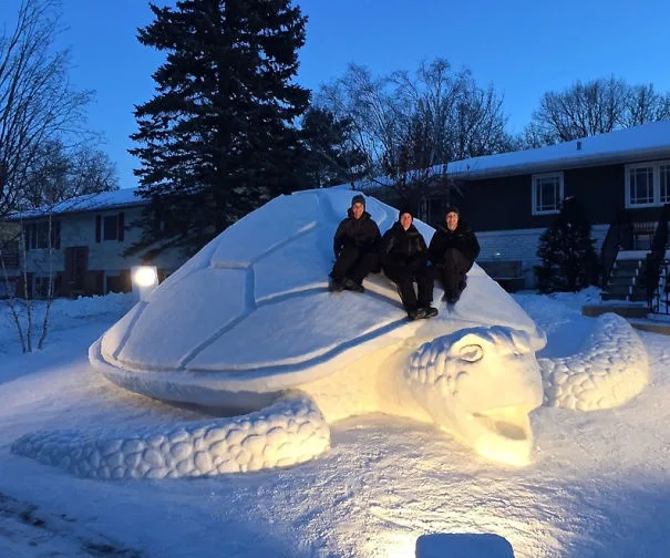 Three people are sitting atop a large, intricately carved snow turtle sculpture in a snowy yard at dusk. The turtle is…