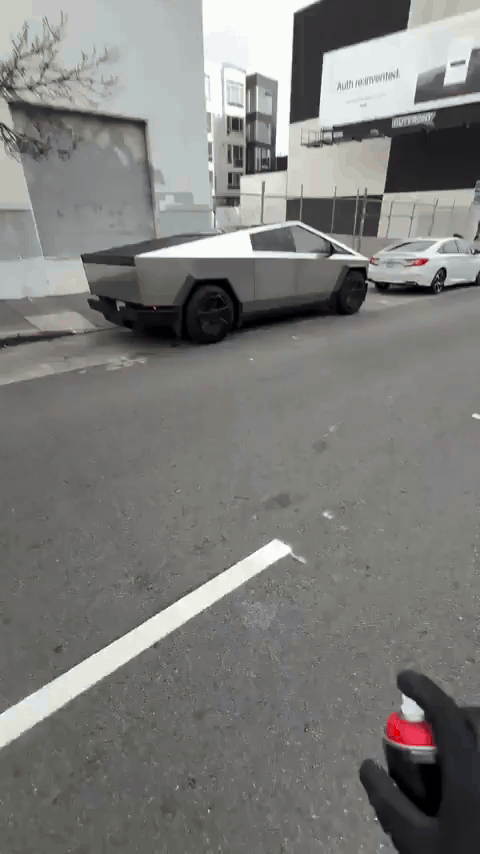 A person wearing black gloves uses red spray paint to draw a crude outline of a car on the side of a silver Tesla Cybertruck…