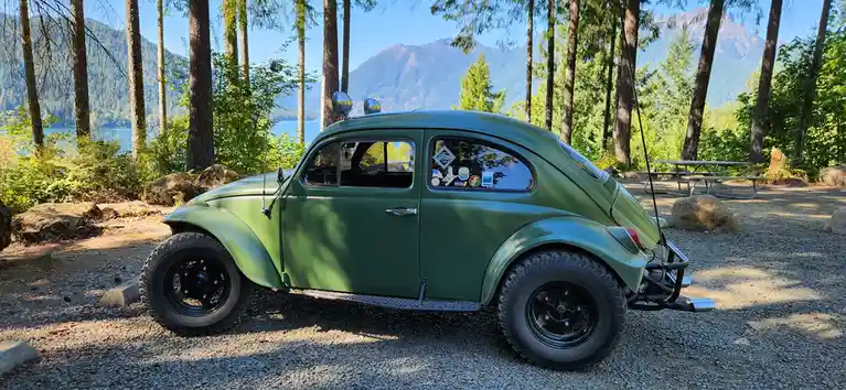 A green vintage Volkswagen Beetle with oversized off-road tires and a front bumper is parked on gravel in a forested…