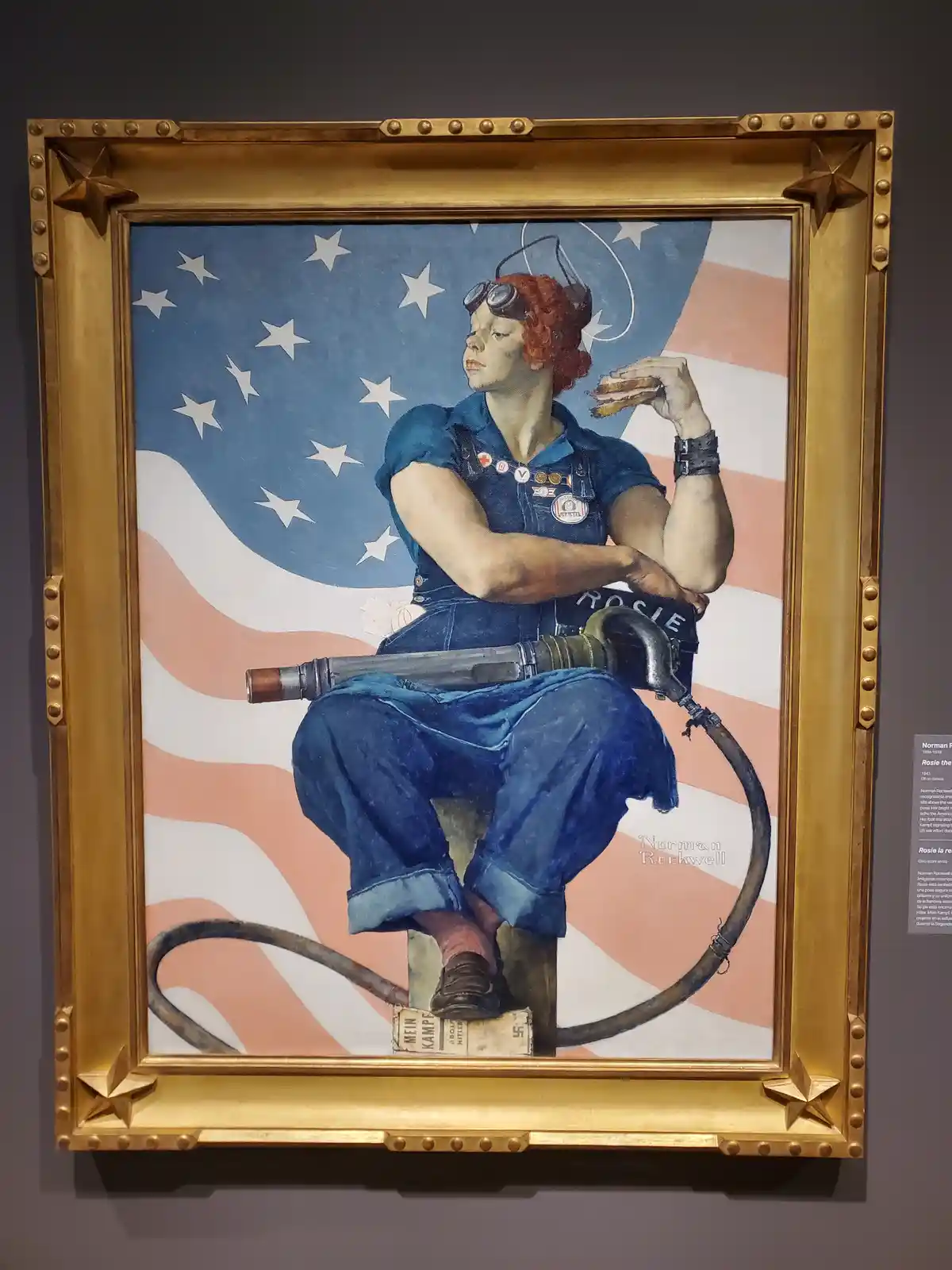 A framed painting of Rosie the Riveter, a woman in blue overalls and goggles, sitting on a stool while eating a sandwich…