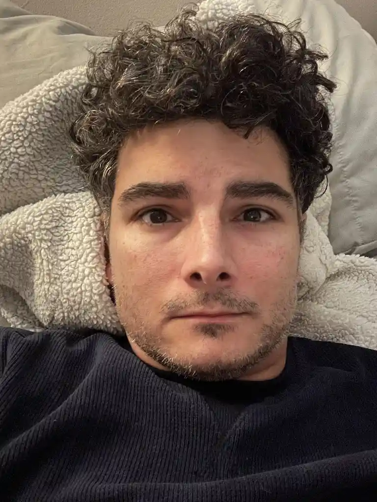 A close-up selfie of a young man with dark curly hair and a light beard, lying on his back on a fluffy white blanket or…