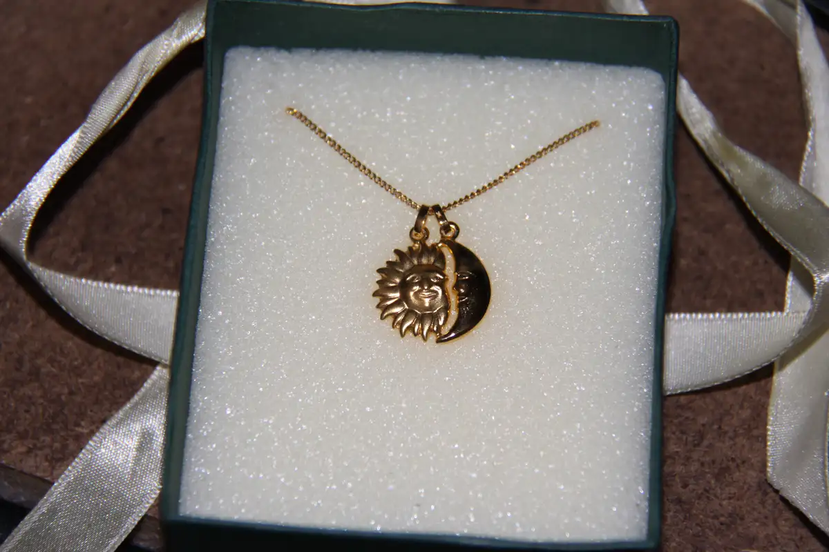 A gold-colored necklace with a sun and moon pendant rests in a glittery white jewelry box lined with a dark green velvet…
