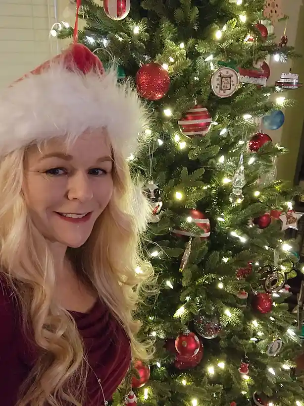 A woman with blonde hair wearing a red and white Santa hat smiles at the camera while standing in front of a decorated…