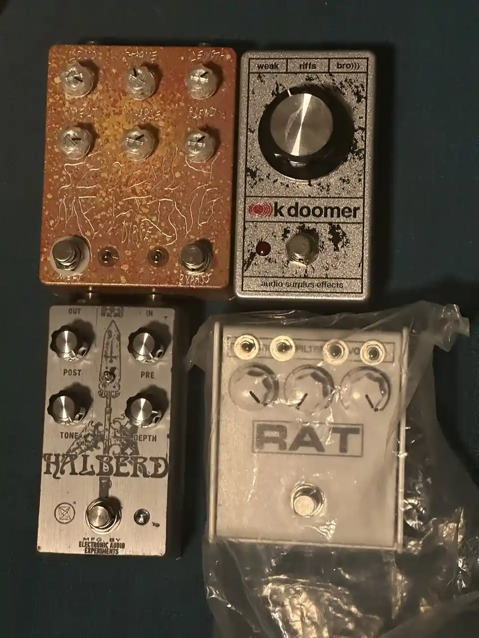 Four guitar effects pedals are arranged on a dark surface. The top-left pedal is a rusted, custom-modified unit with…