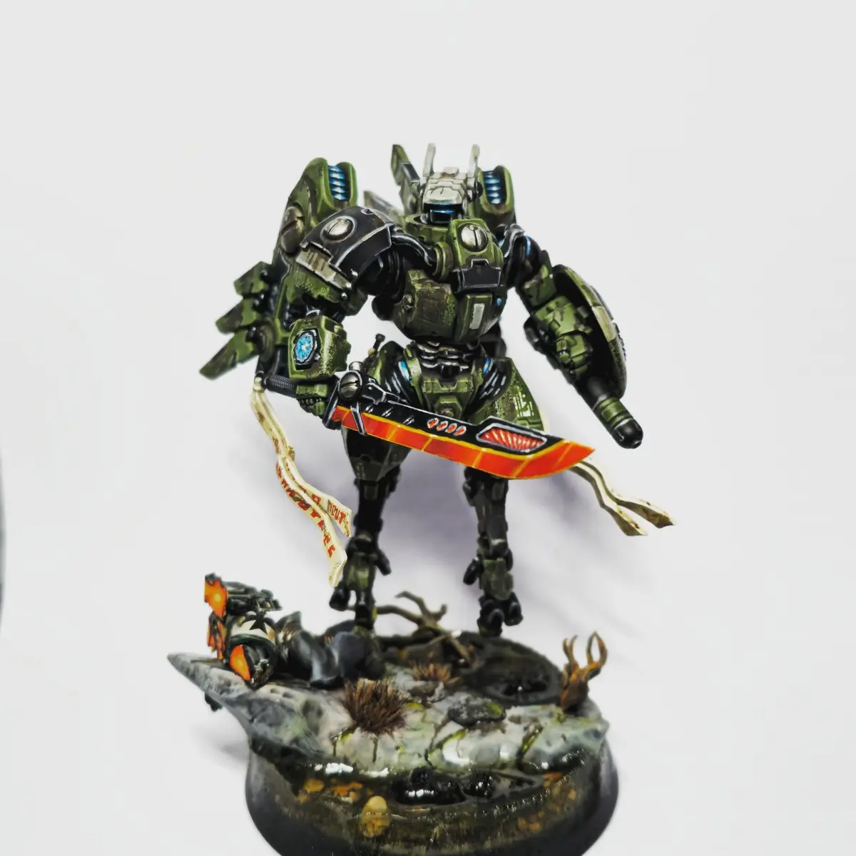 A heavily detailed green and black painted mech miniature stands on a rocky, debris-strewn base, holding a large red-bladed…