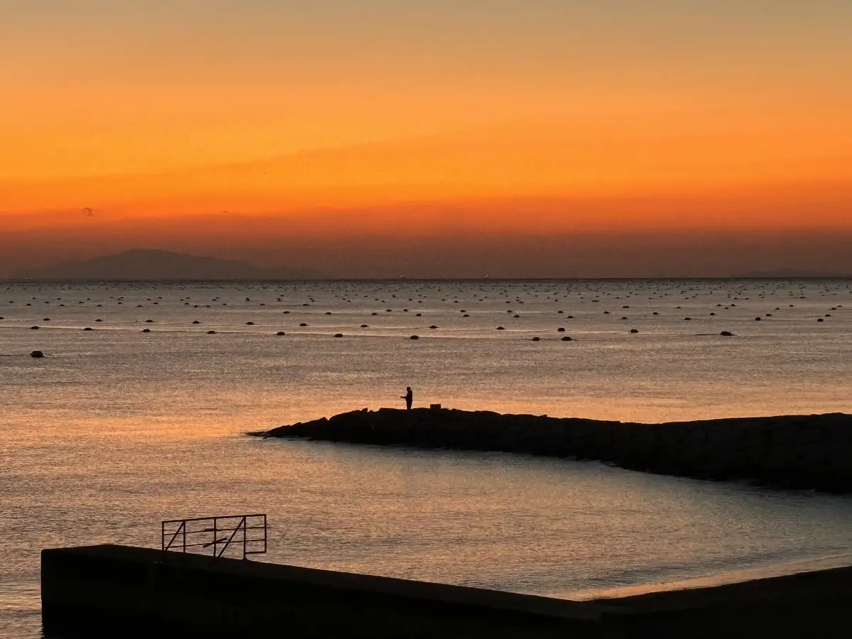 A serene sunset over the ocean with a person silhouetted on a rocky jetty, holding a fishing rod. The sky is a gradient of…