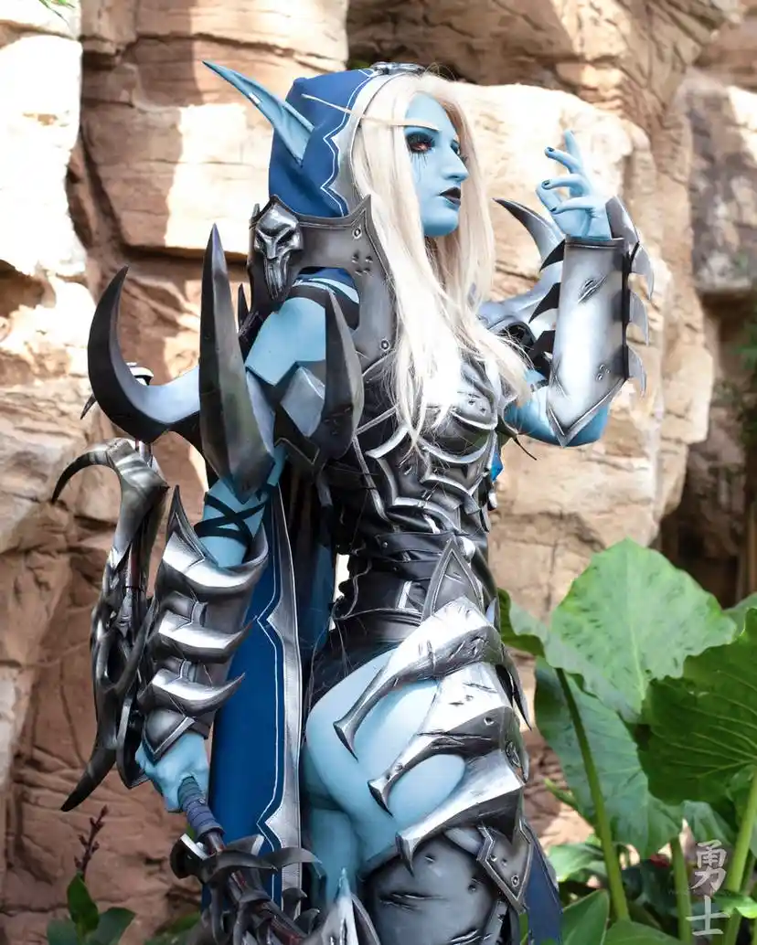 A person in elaborate blue-skinned fantasy armor with pointed ears and ornate silver and black plate armor stands outdoors…