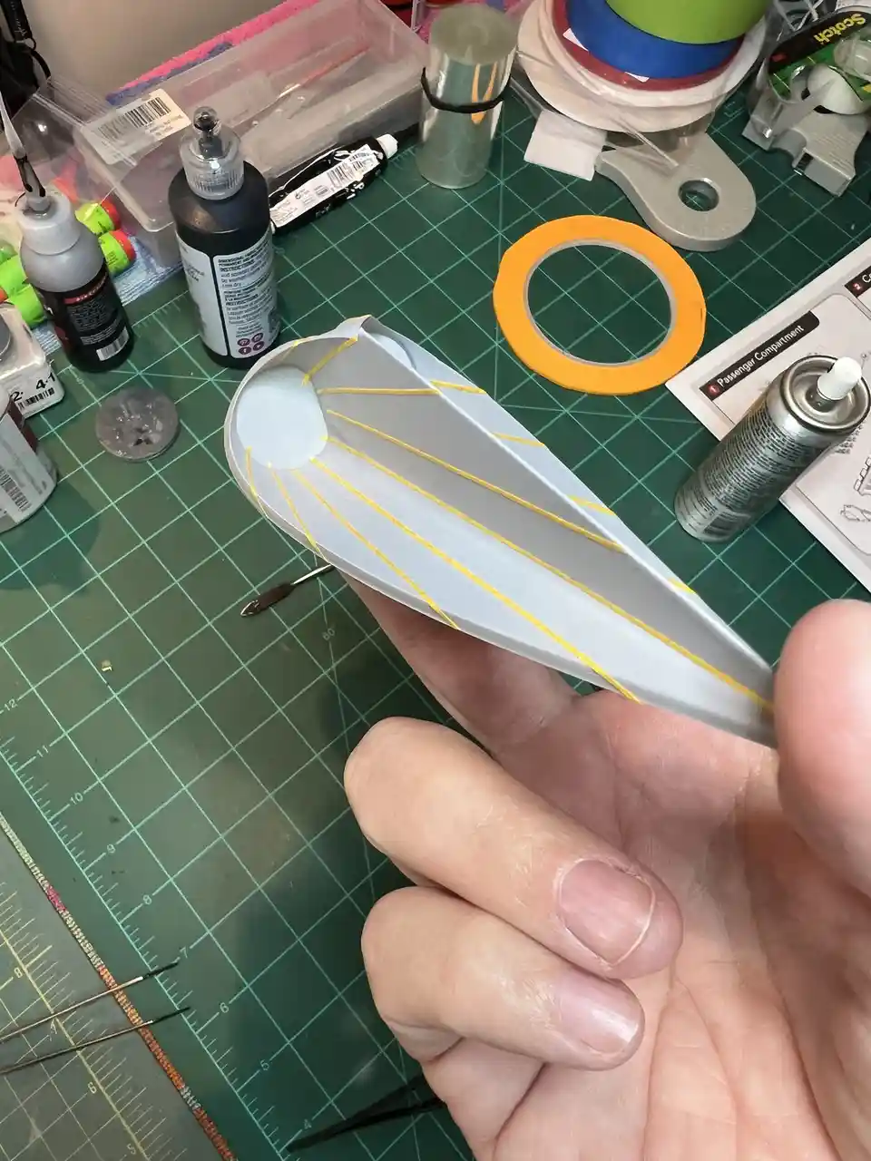 A hand holds a small, gray, partially assembled model airplane fuselage with yellow paint lines radiating from a white…