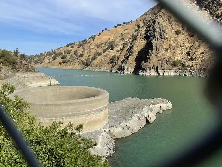 A photograph of a reservoir with visibly low water levels, showing exposed concrete structures and dry, brown hillsides…