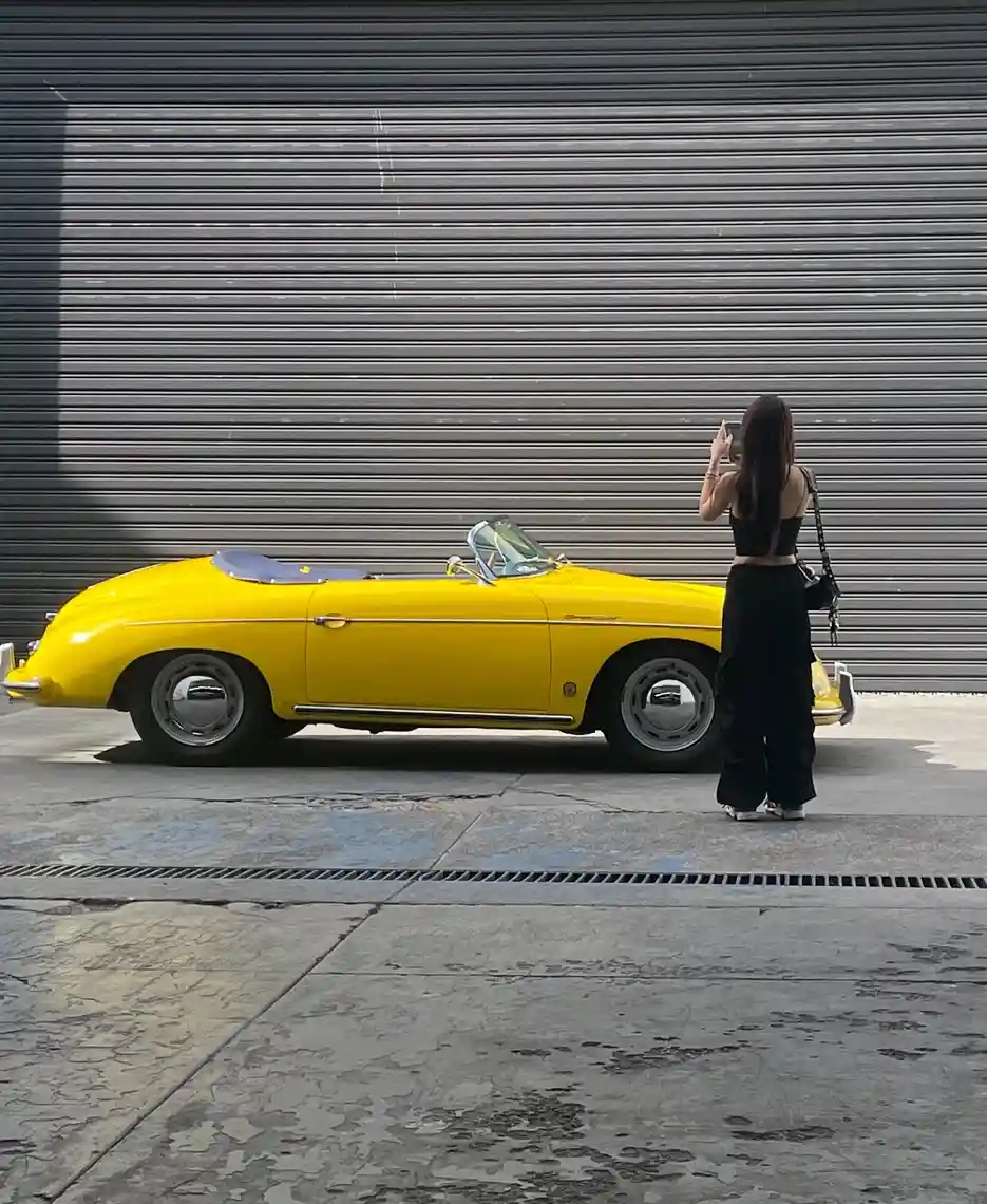 A person with long dark hair, wearing a black crop top and black pants, stands beside a bright yellow vintage convertible…
