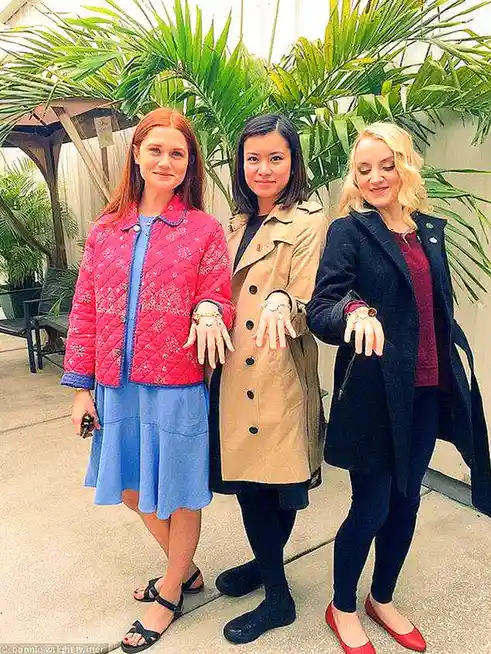 Three women stand side-by-side outdoors, each holding up their hands to display multiple rings on their fingers. The woman…