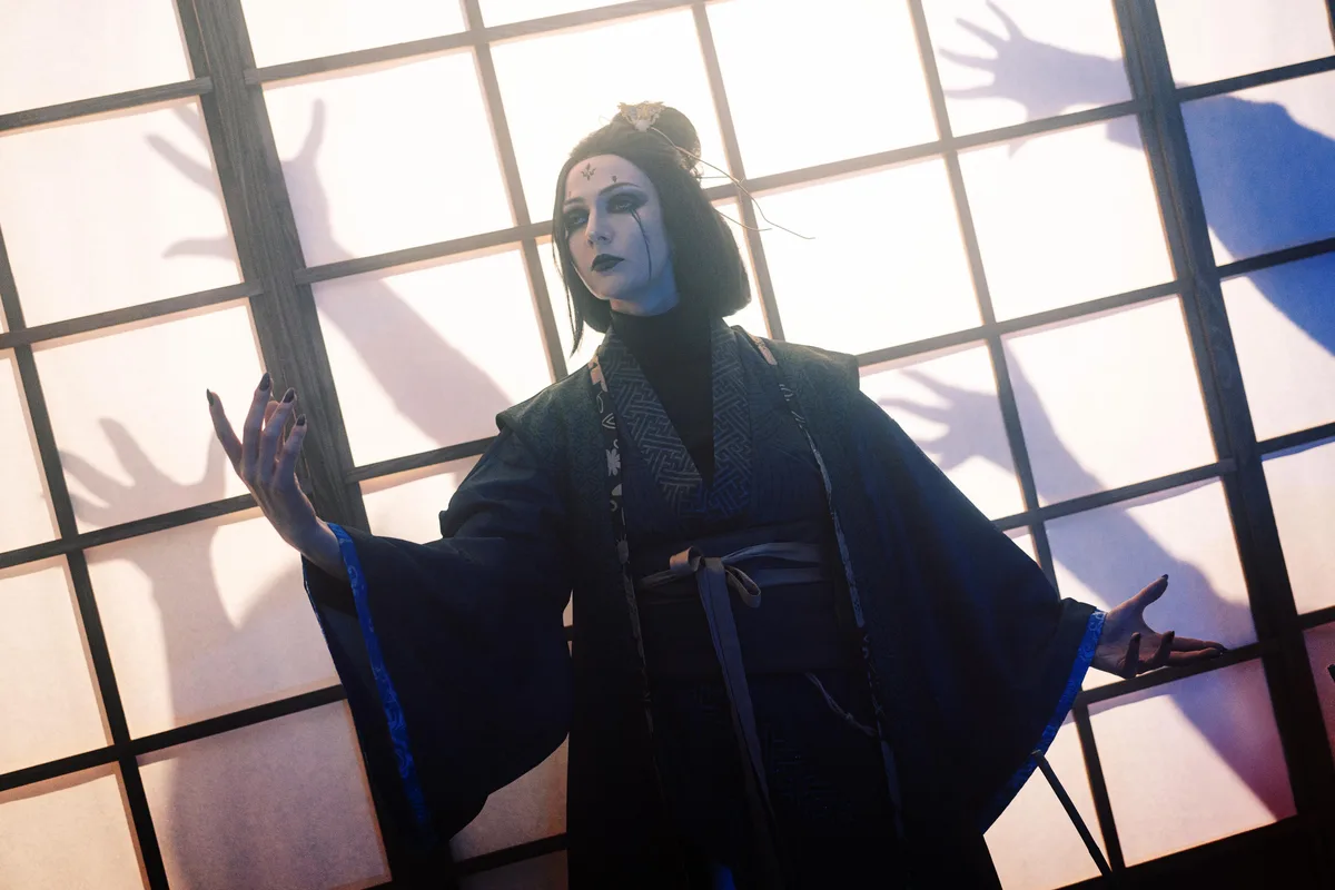 A person in dark traditional Japanese attire with dramatic makeup and black nail polish stands with arms outstretched…