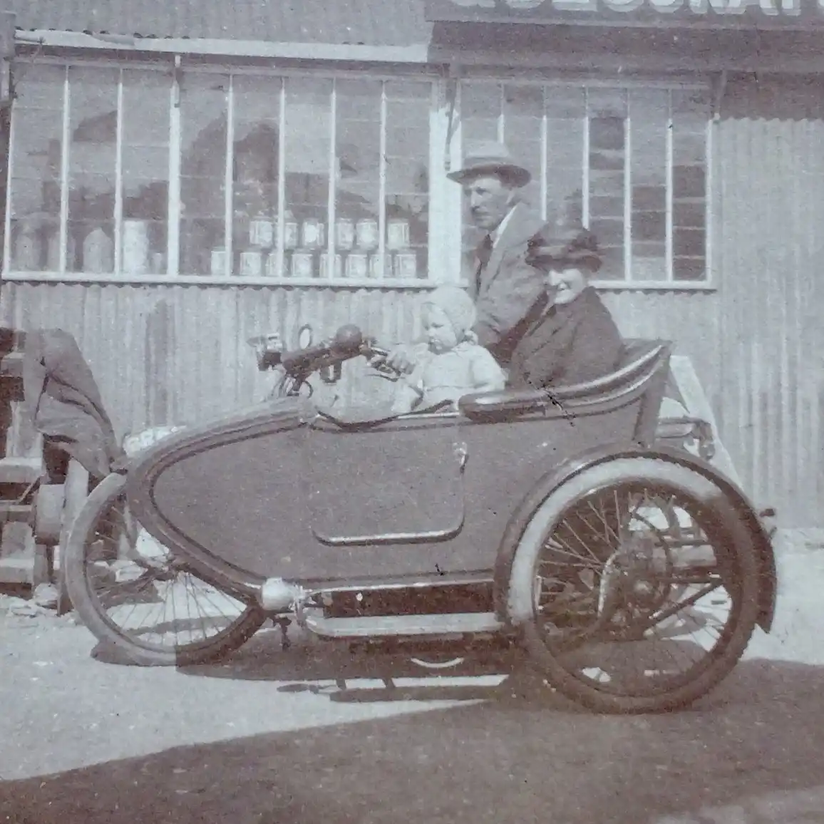 A vintage black-and-white photo shows a baby in a small, old-fashioned three-wheeled motorized vehicle, gripping the…
