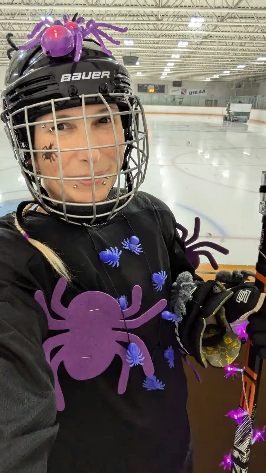 A person in a black Bauer hockey helmet and jersey stands on an ice rink, decorated with purple and blue plastic spiders on…
