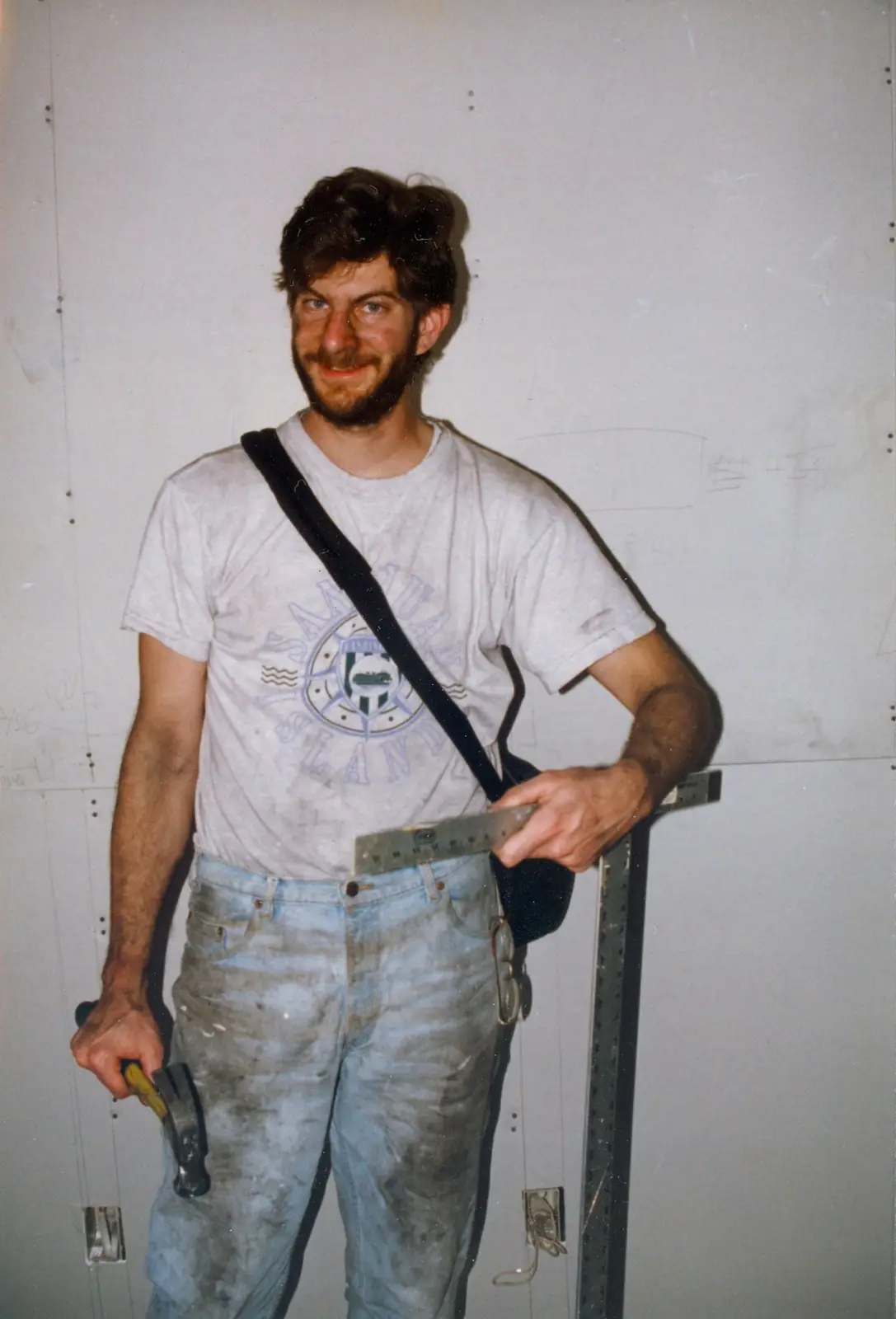 A man with a beard and messy hair stands in front of a drywall wall, holding a hammer in one hand and a level in the other…