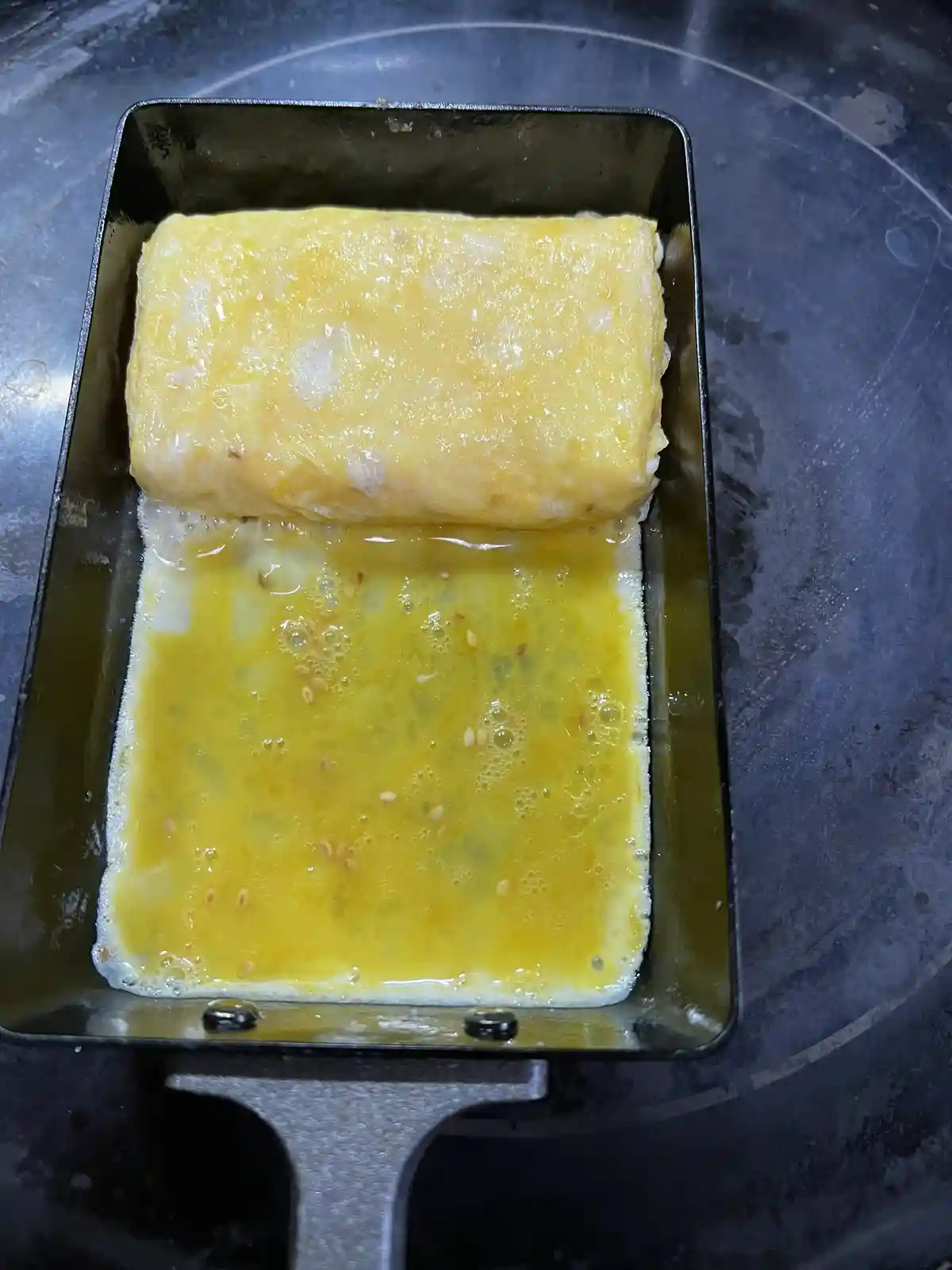 A rectangular metal pan contains a partially cooked, yellow egg omelette. The bottom layer is still liquid egg with sesame…