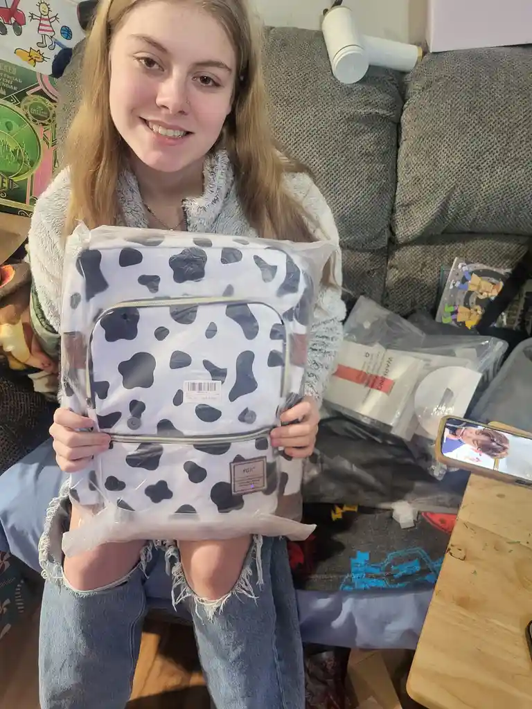 A young woman with long blonde hair, wearing a fuzzy grey top and ripped jeans, sits on a couch holding a new cow-print…
