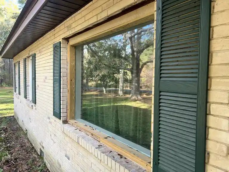 A photo of a house with light-colored brick walls and dark green shutters, showing a window with a new wooden frame and…