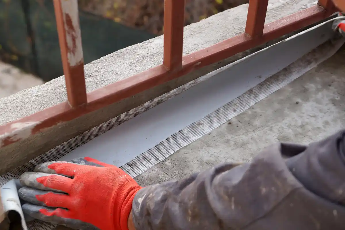 A close-up shot shows a person wearing a red and grey work glove pressing down a white mesh-backed strip along a concrete…