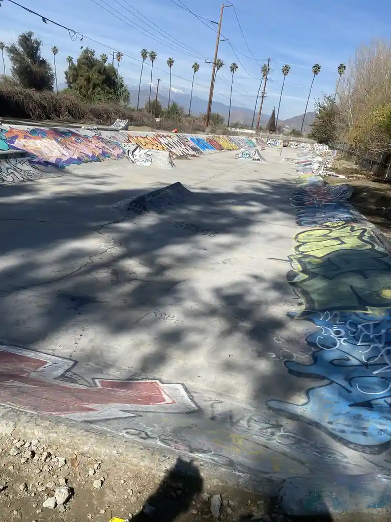 An outdoor concrete skatepark covered in colorful graffiti, with ramps and bowls under a sunny sky with palm trees and…