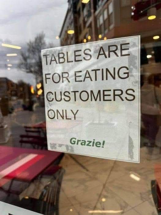 A sign taped to a restaurant window reads 'TABLES ARE FOR EATING CUSTOMERS ONLY' in bold black text, with 'Grazie!' in green…