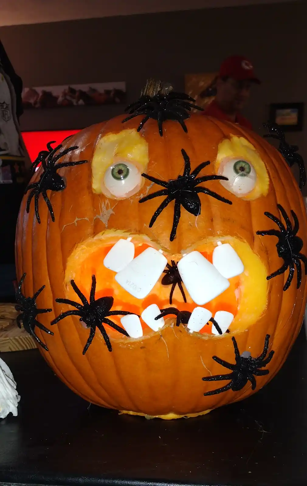 A carved orange pumpkin with large green plastic eyes, a wide mouth filled with white marshmallows as teeth, and numerous…
