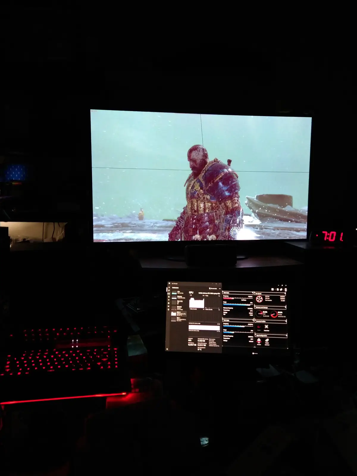 A dark room with a large monitor displaying a snowy scene from a video game featuring a heavily armored, bloodied warrior…