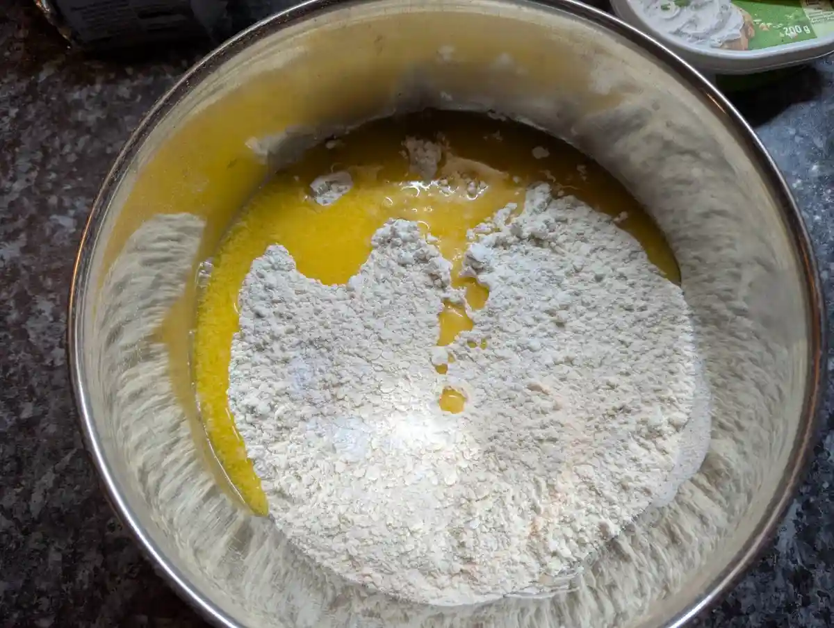 A top-down view of a metal mixing bowl containing a pool of yellow liquid, likely beaten eggs or melted butter, with a large…