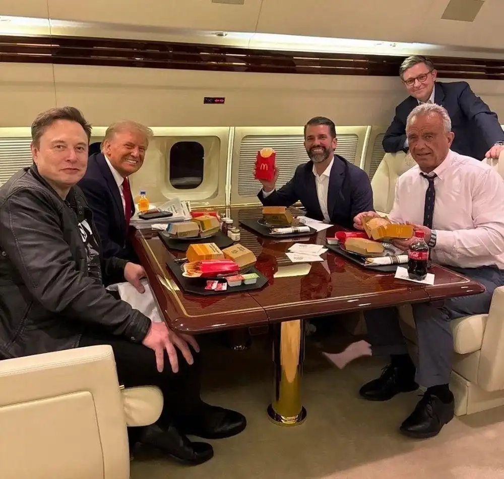 Four men are seated around a polished wooden table inside a private jet cabin, eating McDonald's food from boxes and cups…