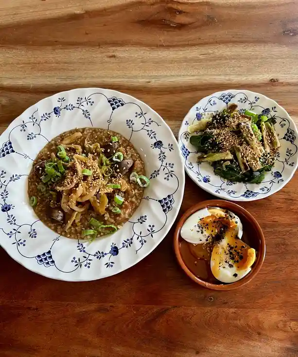An overhead photo shows three dishes on a wooden table: a large bowl of savory rice topped with mushrooms, green onions, and…