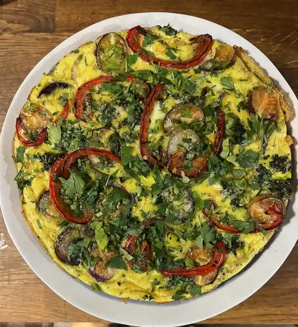 A top-down photo of a round, yellow egg-based dish on a white plate, resembling a pizza with toppings like red bell peppers…