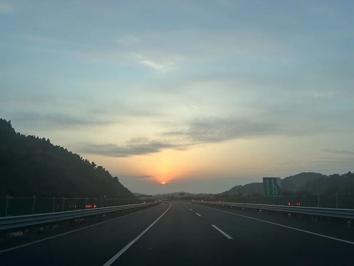 A photo taken from a car on a highway at sunset, showing a wide road with lane markings, guardrails, and distant hills under…