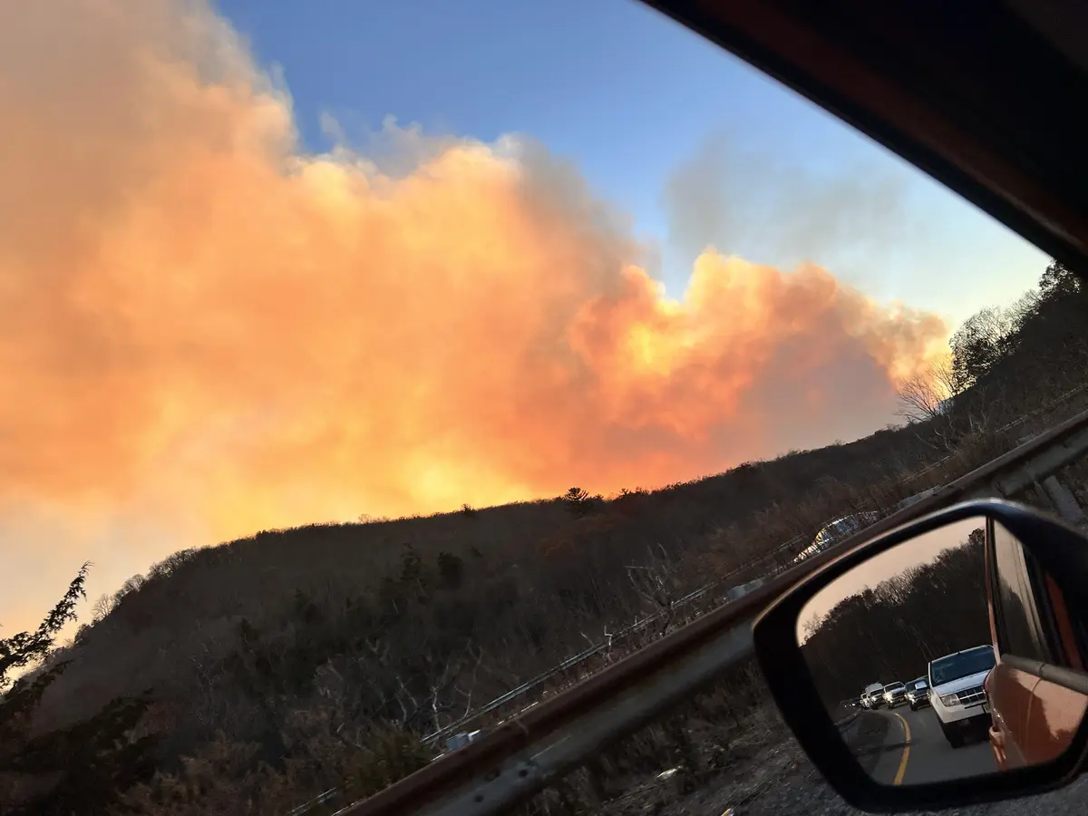 A photo taken from inside a car shows a large, orange-hued smoke plume rising from a forested hillside under a blue sky. The…