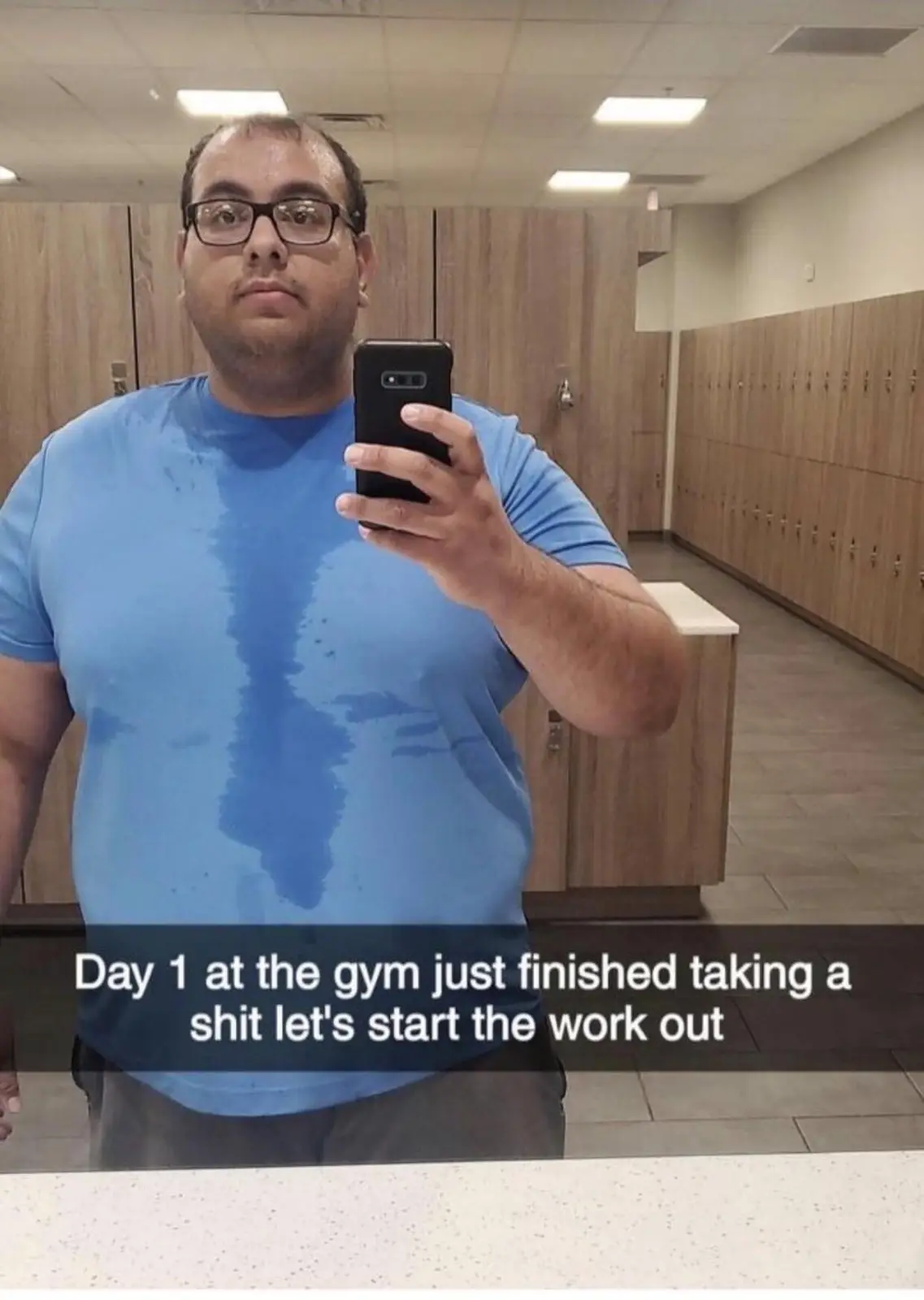 A man in a blue t-shirt with a large sweat stain down the front takes a mirror selfie in a gym locker room, holding a black…