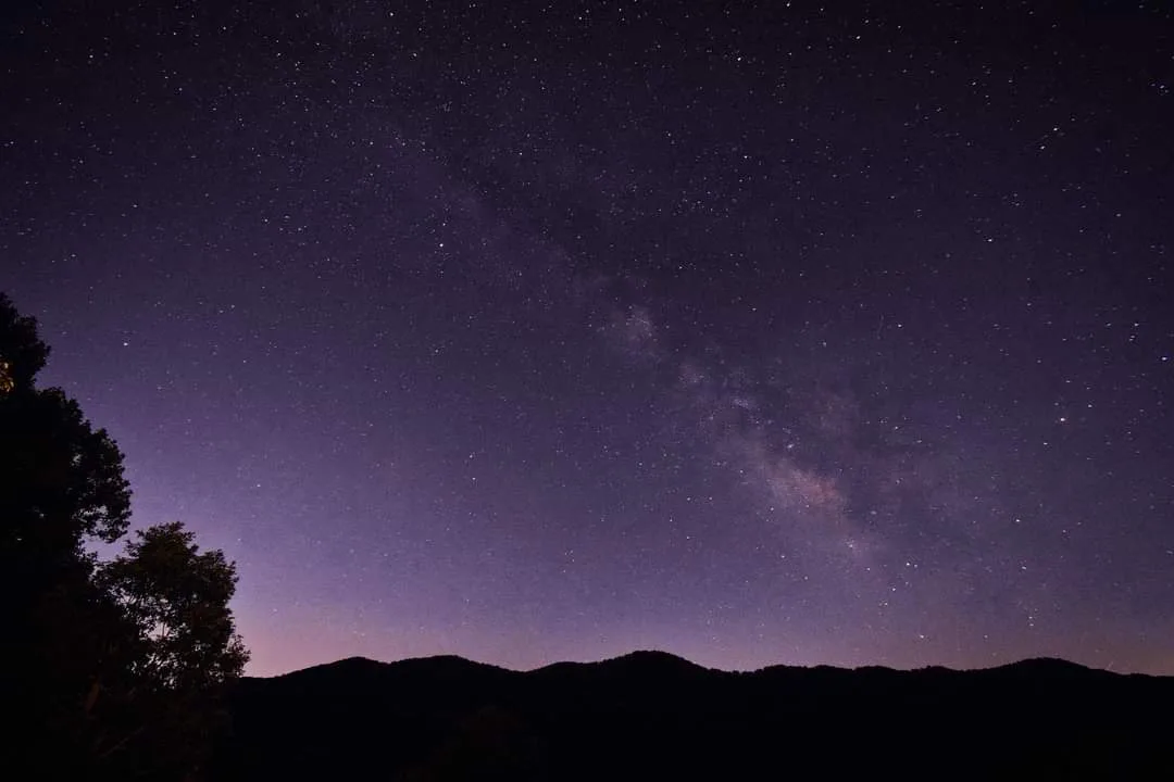 A stunning night sky photograph showing the Milky Way galaxy as a bright, cloudy band stretching across a dark purple sky…