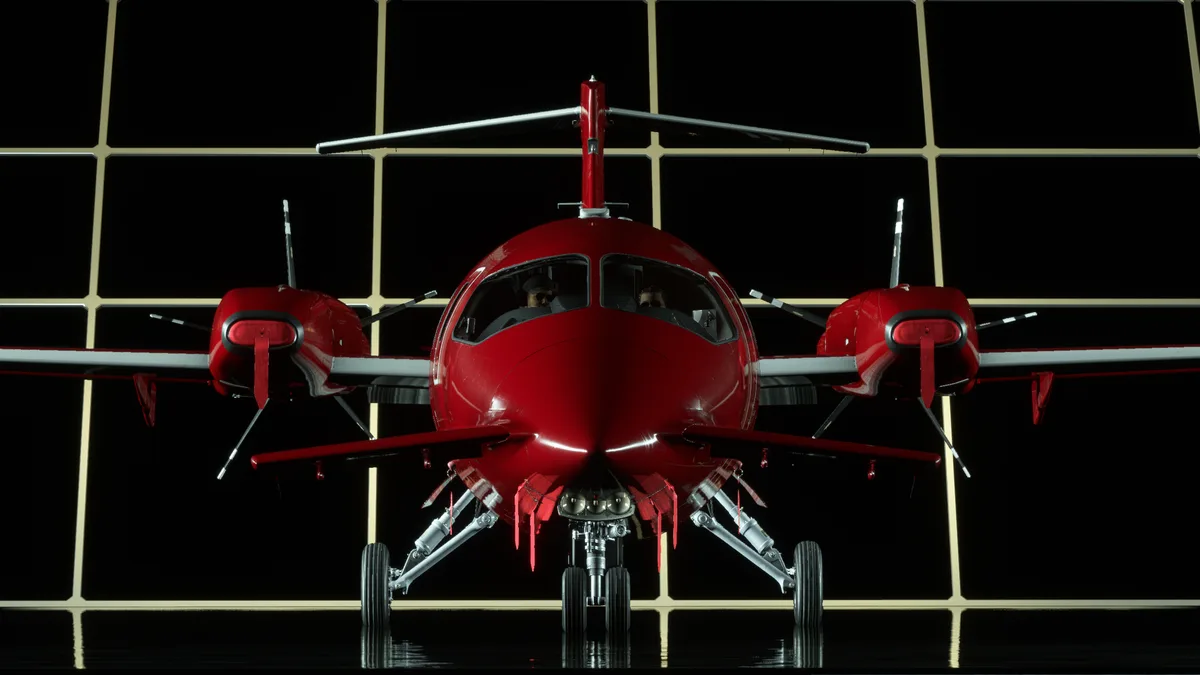 A glossy red twin-engine propeller airplane is shown head-on, parked on a reflective black floor in front of a grid of black…