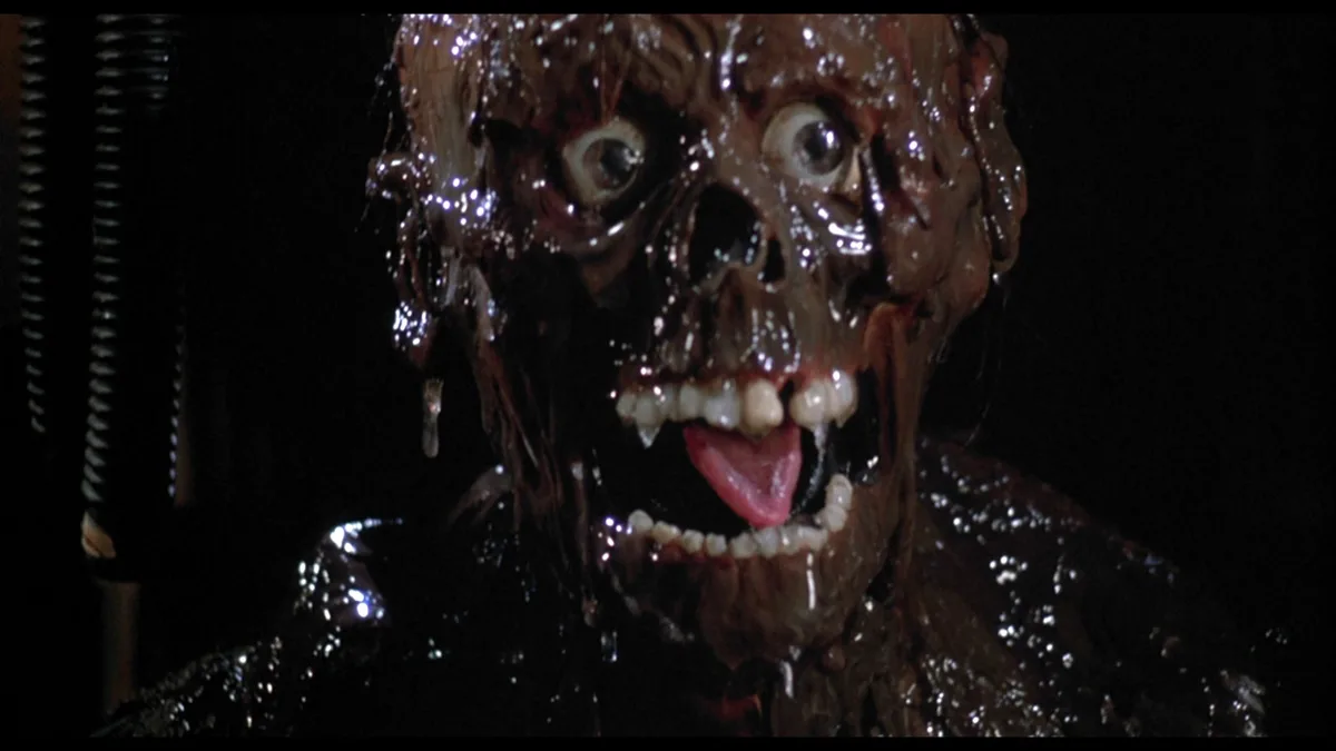 A close-up shot of a grotesque, melted, and oozing humanoid figure with exposed teeth and a tongue, covered in dark, viscous…