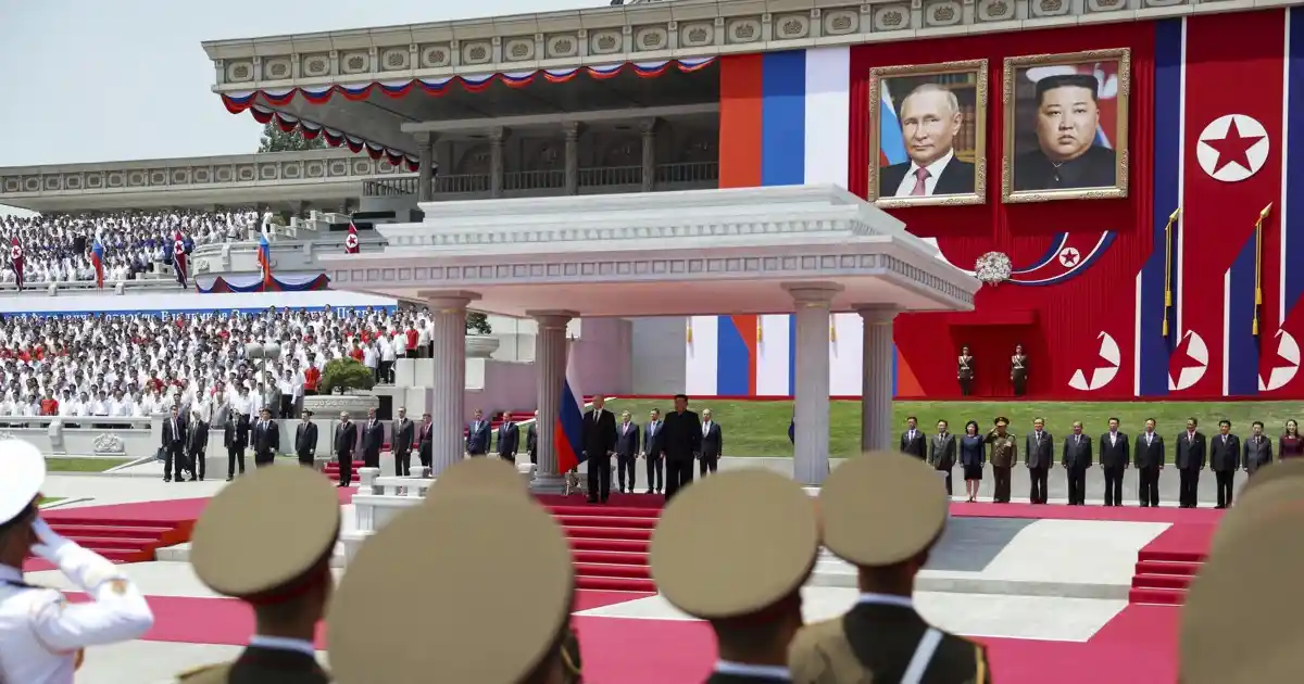 A large outdoor ceremony with many people in formal attire, framed by red carpets and flags. Two large portraits hang…