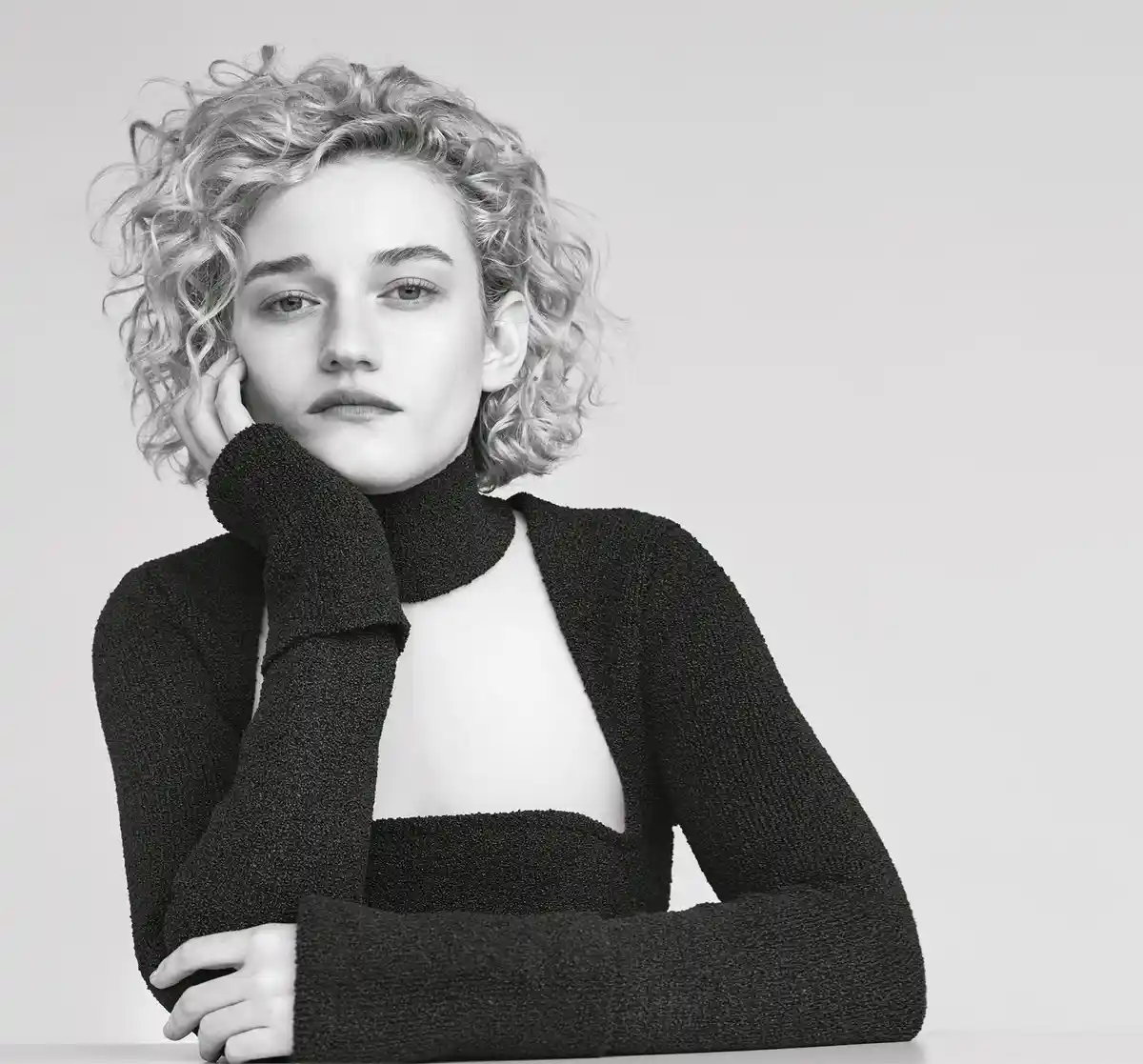 A black-and-white portrait of a woman with curly blonde hair, wearing a black textured turtleneck top with a cutout at the…