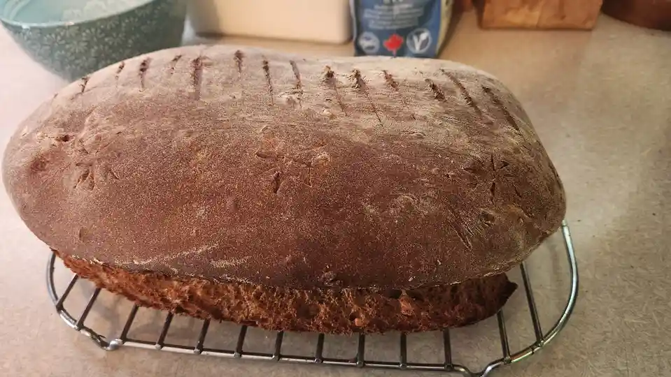 A freshly baked, dark brown, oval-shaped loaf of bread with scoring marks sits on a metal cooling rack. The bread has a…