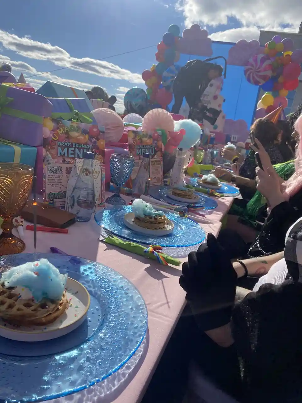 A long outdoor table is set for a drag brunch with bright blue plates, waffles topped with blue cotton candy, rainbow…