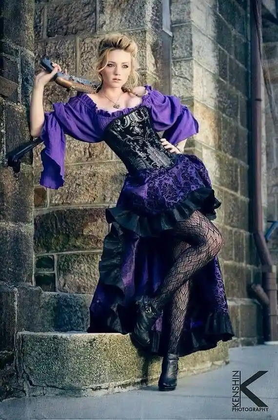 A woman in a purple and black gothic-style dress with puffy sleeves, fishnet stockings, and a corset, poses on stone steps…