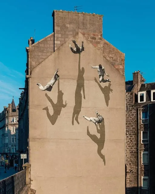 A large mural on a building facade depicts four figures in mid-air, seemingly falling or leaping, with their elongated…