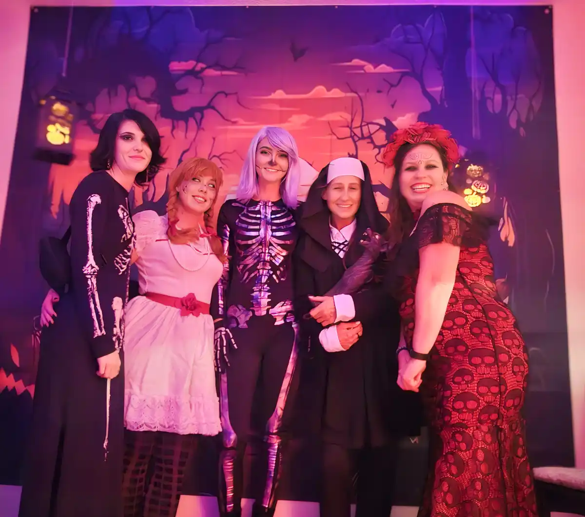 Five women pose together in Halloween costumes against a spooky backdrop with bare trees and bats. From left to right: one…