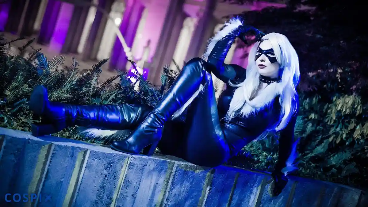 A person in a black leather Catwoman costume with white fur trim and a black mask poses sitting on a low wall at night…